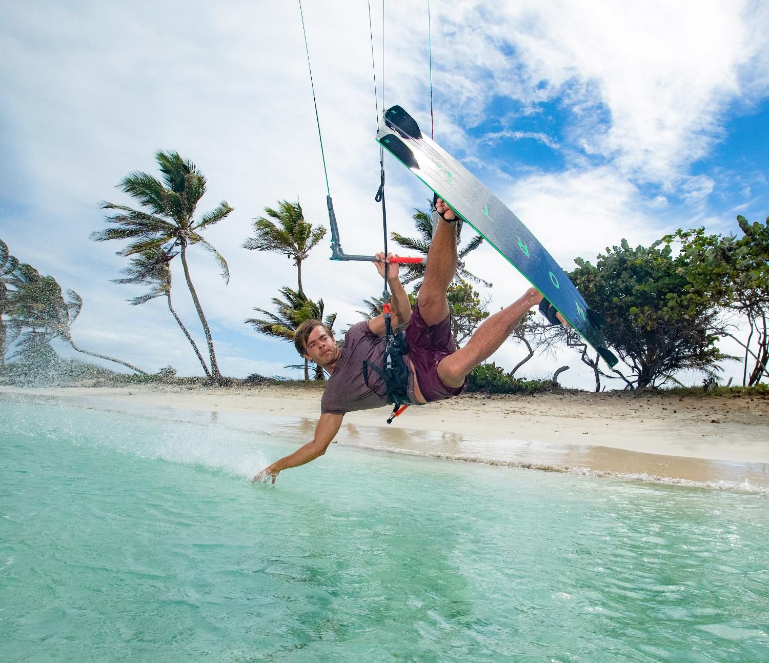 Kitesurfer performing a trick over clear water with palm trees in the background.