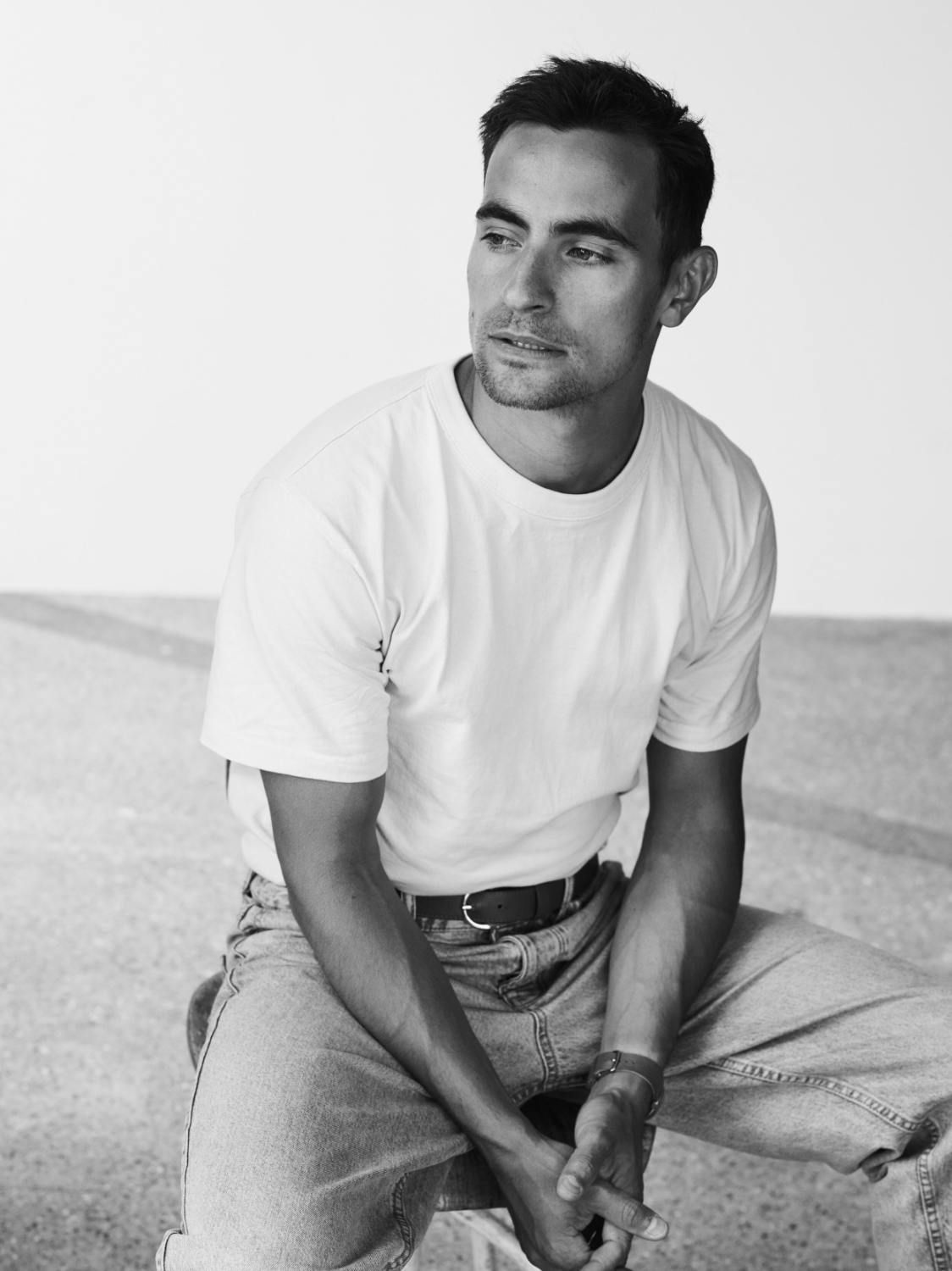 Jonathan Harboe sits casually, wearing a white t-shirt and jeans, with a thoughtful expression.