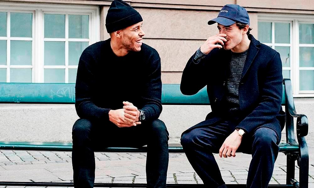 Mathias Zanka and another man seated on a bench, sharing a lighthearted conversation.