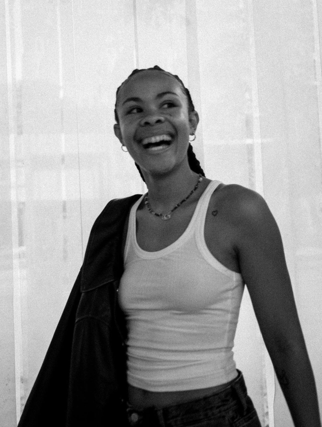 Bathsheba smiles warmly, wearing a white tank top and holding a jacket, with sheer curtains behind.