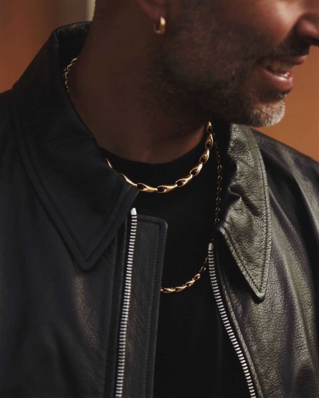 Mads Emil Møller wearing a black leather jacket and gold jewelry, smiling softly.