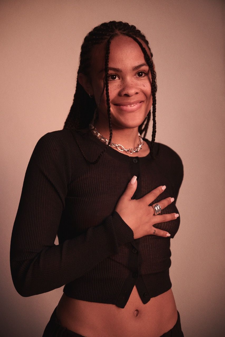 Bathsheba smiles warmly, wearing a black top and layered jewelry against a soft background.