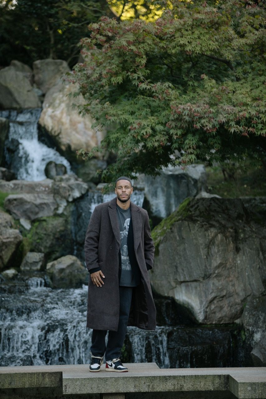 Man in a long coat stands by a waterfall surrounded by greenery and rocks.