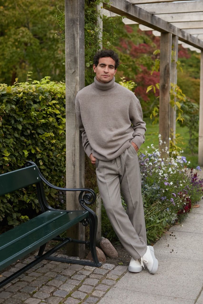 Jonathan Harboe poses casually in a garden, wearing a sweater and trousers, with greenery around.
