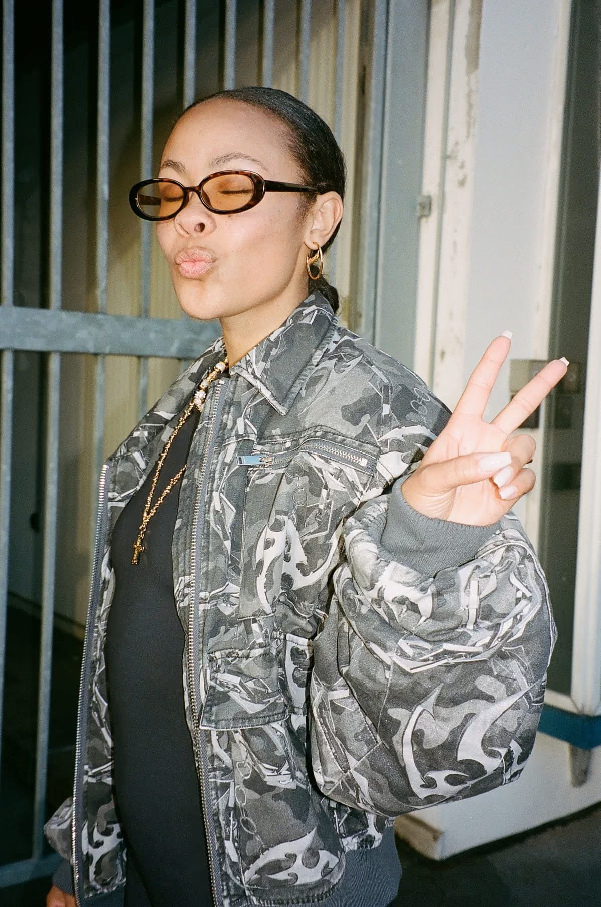 Bathsheba poses playfully in a camouflage jacket, making a peace sign against a backdrop.