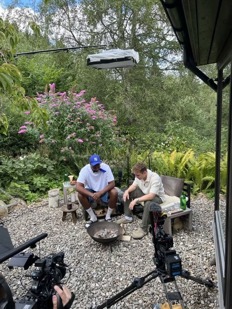 Rasmus Brohave and another individual sit by a fire pit in a lush outdoor setting.